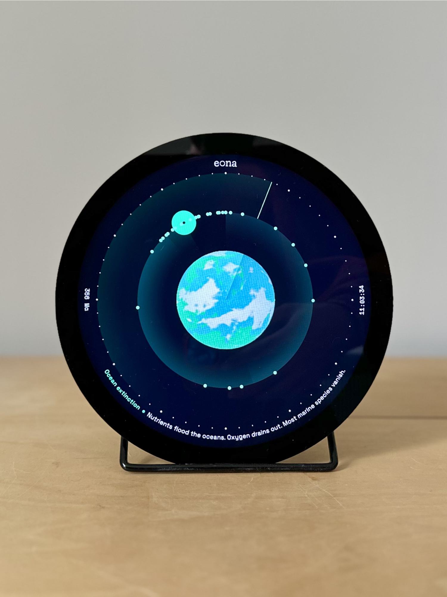 The physical eona device, a round display on a stand showing the Ocean extinction milestone, standing on a wooden desk